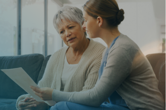 Hausbesuch einer Mitarbeiterin des HypoPT Ascendis Care Betreuungsprogramms bei einer Patientin mit HypoPT. Sie sitzen zusammen auf der Couch – die Frau zeigt der Patientin Infomaterialien.
