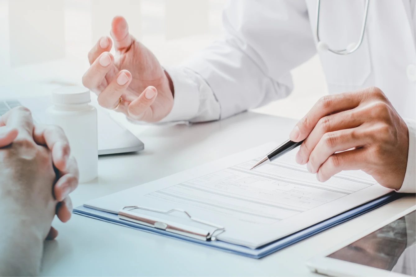 Ärztin oder Arzt im Patientengespräch. Der Bildausschnitt zeigt die Hände von zwei Personen, die sich am Tisch gegenüber sitzen. Vor der Ärztin oder dem Arzt liegen ein Klemmbrett und ein Tablet.