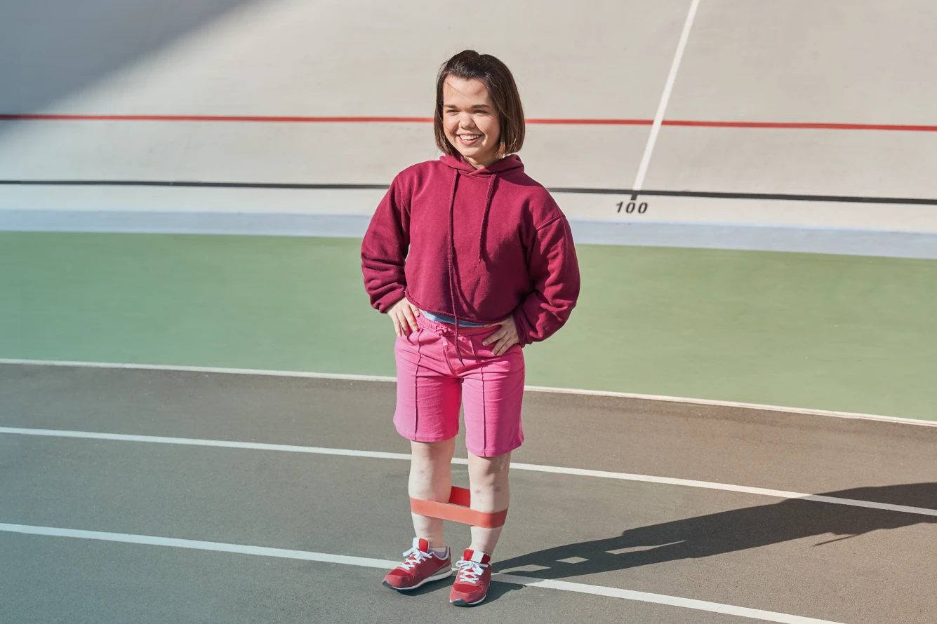 Selbstbewusst lächelnde kleinwüchsige Frau trainiert mit einem Resistance-Band auf der Laufbahn eines Stadions.