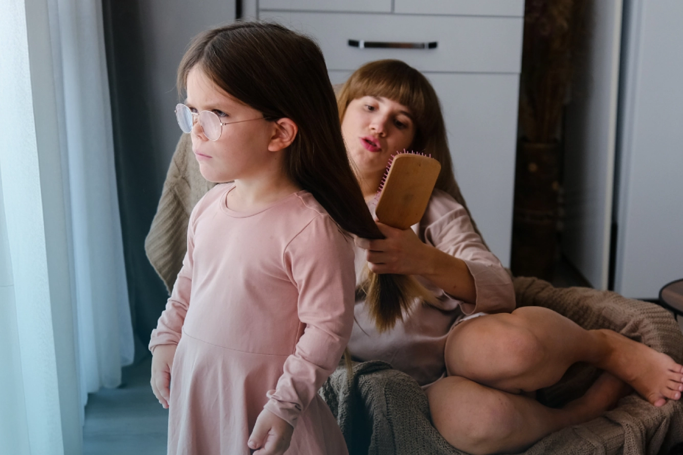 Mutter sitzt zu Hause im Sessel und bürstet ihrem kleinwüchsigen Kind die Haare; das Kind steht vor ihr und schaut aus dem Fenster.