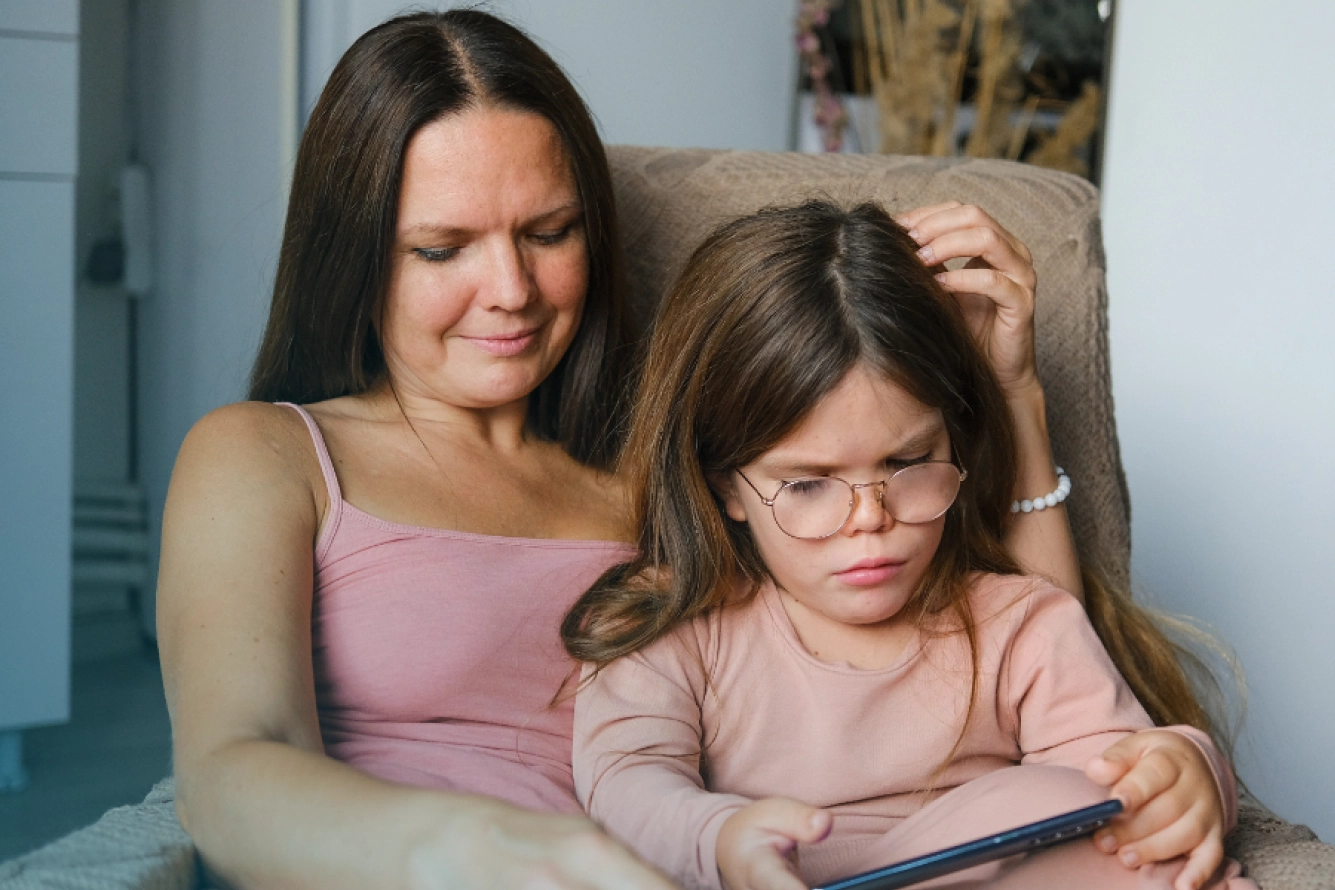 Mutter sitzt mit ihrer kleinwüchsigen Tochter auf dem Schoß in einem gemütlichen Sessel und schaut ihr beschützend beim Lesen zu.